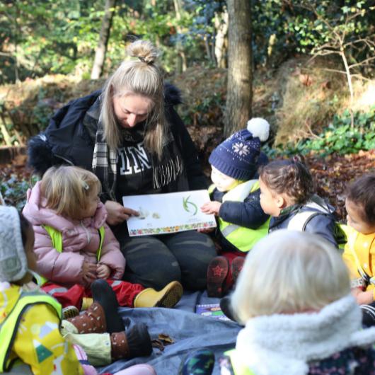 staff reading a book to a group of children sitting outside in the local park