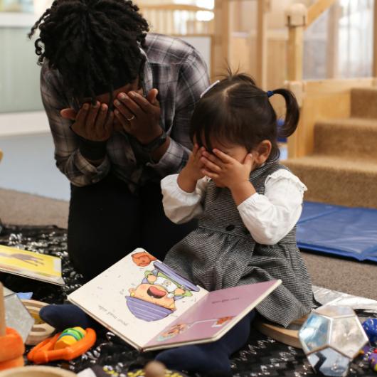 Nursery teacher and child covering their eyes with their hands whilst reading a book