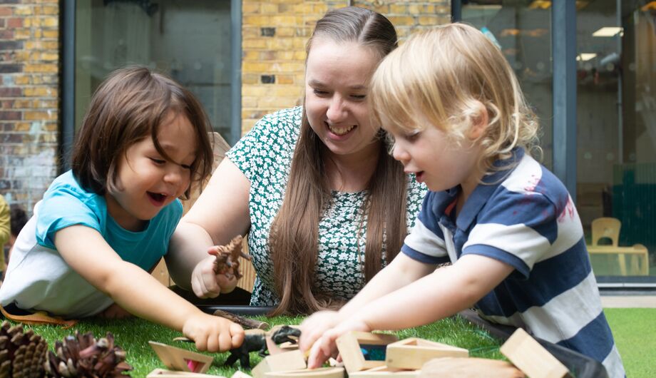 A teacher and children are playing with dinosaur toys and wooden shapes in the garden.