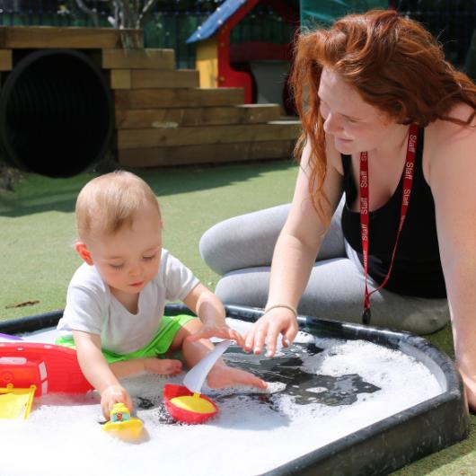 Baby and nursery teacher play in water pit in garden