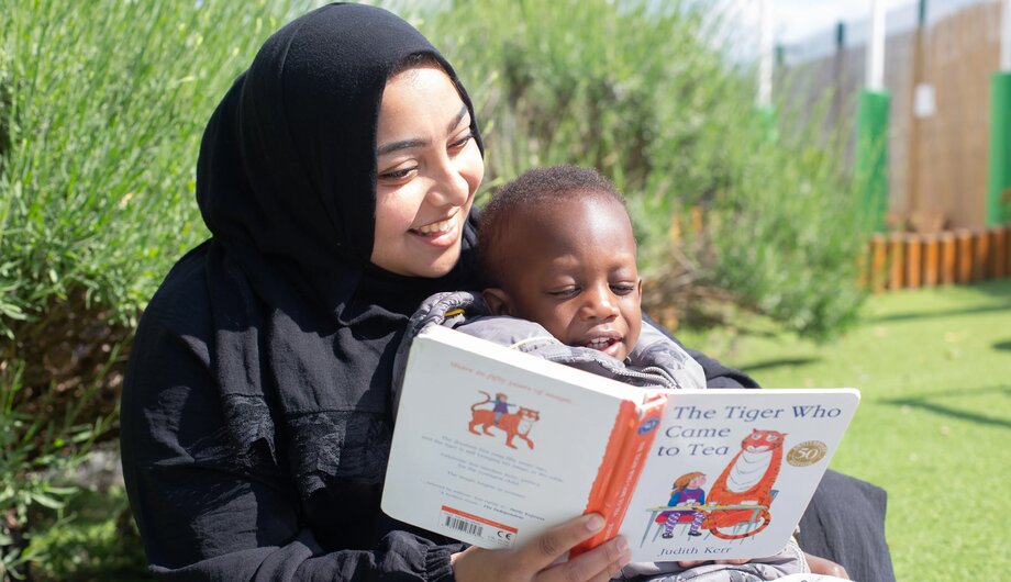 Staff member and child sitting outside reading a book in the sunshine