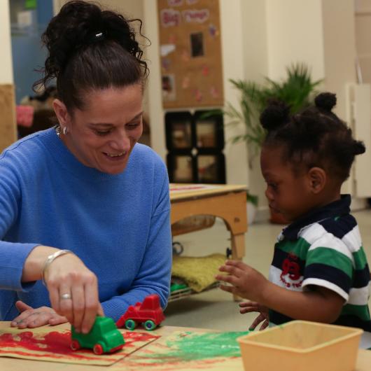 Staff showing child how to paint using the wheels of a plastic car