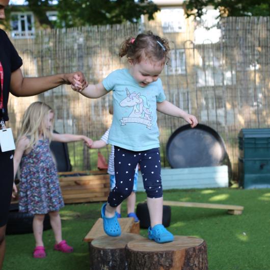 Staff helping a child walk across some wooden logs to practice risky play outside in the garden