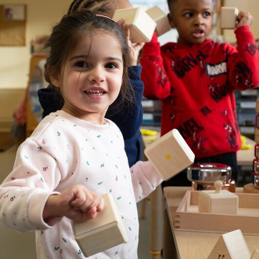 A child is holding wooden block and smiling at teacher