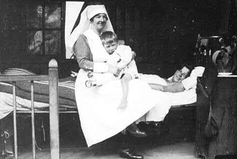 Black and white photo of a nurse holding a child on her lap, sitting on the bed of a sick patient