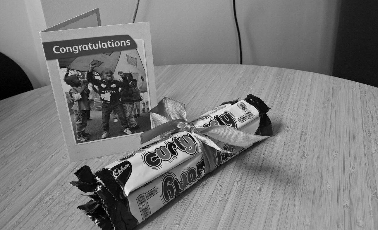 Black and white photo of a Congratulations card next to a bundle of Curly Wurly chocolate bars tied with a ribbon