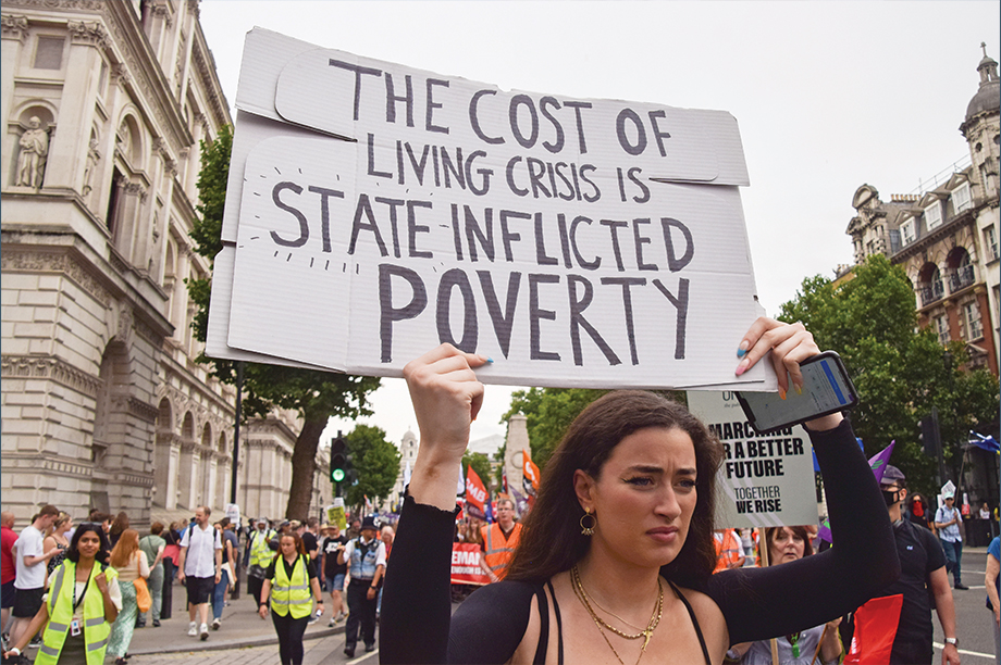 Protesting woman holding a placard stating The Cost of Living Crisis is State Inflicted Poverty