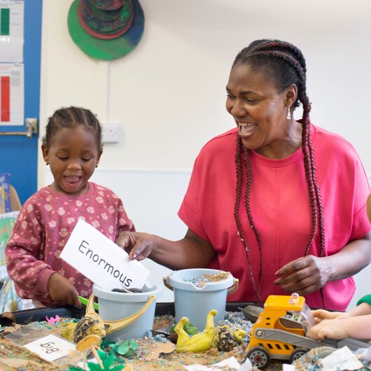 Teacher and children playing in a sandpit with cards with descriptive words 'enormous' and 'big'