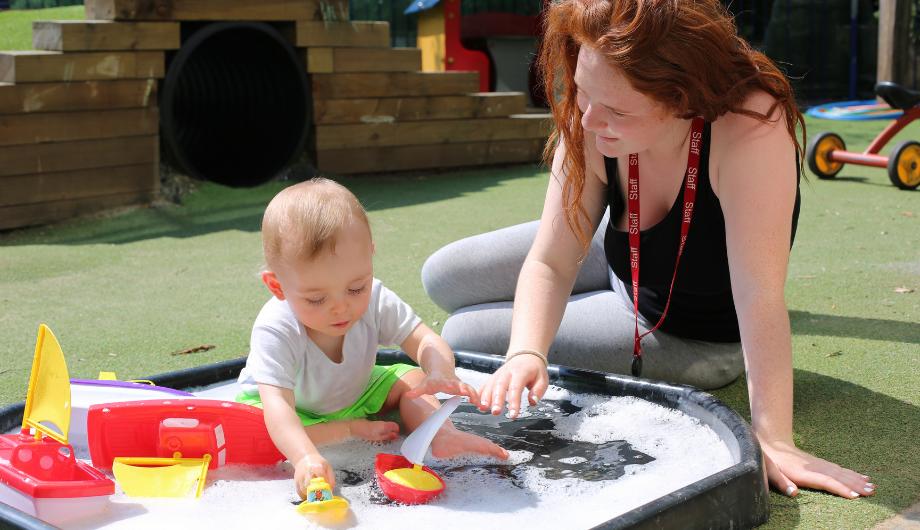 Baby and nursery teacher play in water pit in garden