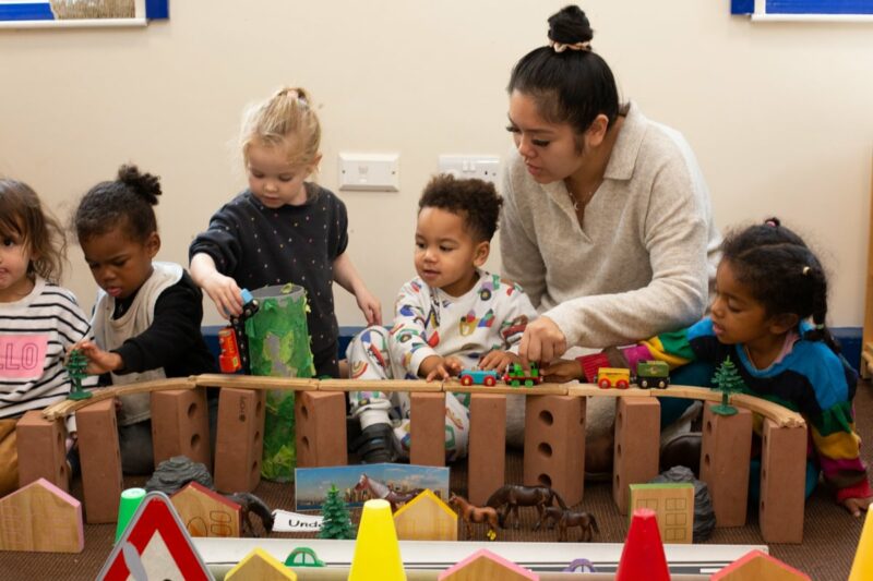 A group of children and a teacher working together to play with trains on a toy wooden bridge
