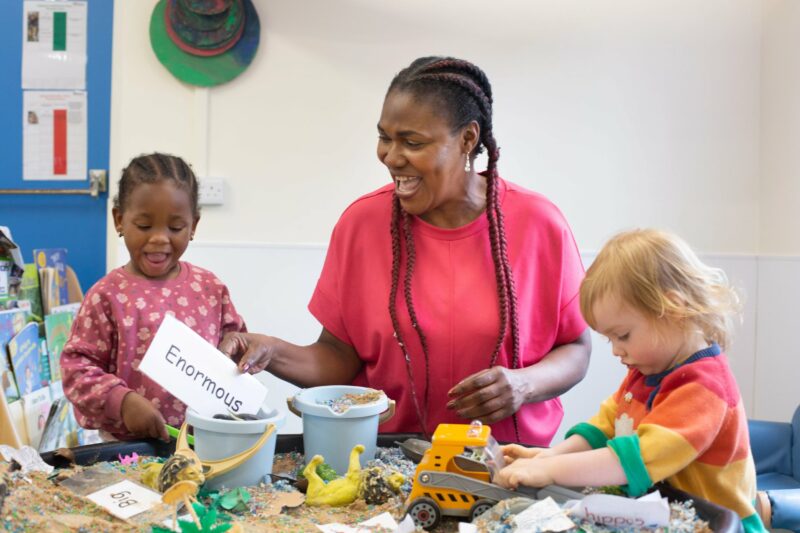 Teacher and children playing in a sandpit with cards with descriptive words 'enormous' and 'big'